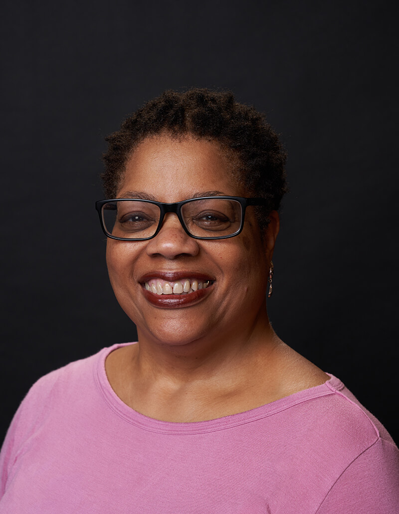 Headshot of Renee Garner Elms, voice actor. She is wearing a pink shirt and is in front of a dark grey background.